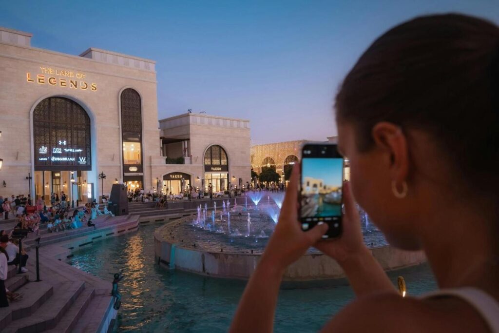 Land of Legends Shopping Avenue illuminated at night with palm trees, shops and visitors in Belek, Antalya